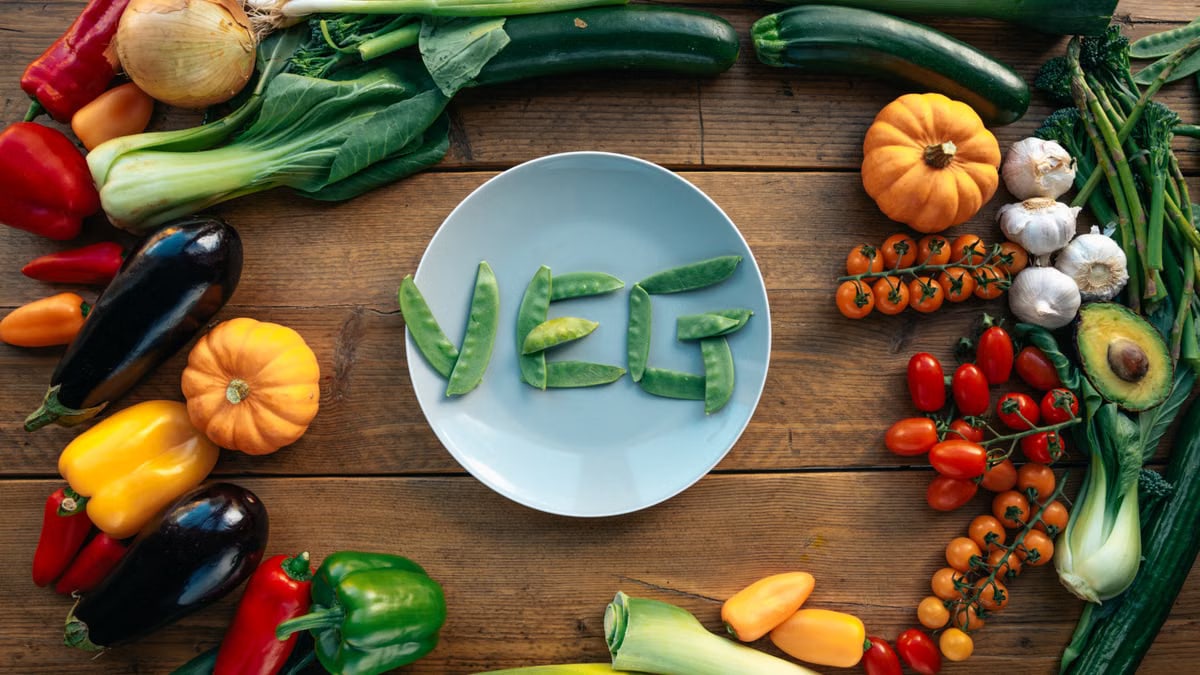 A top view of assorted fresh vegetables arranged around a plate with 'veg' spelled out using peas.